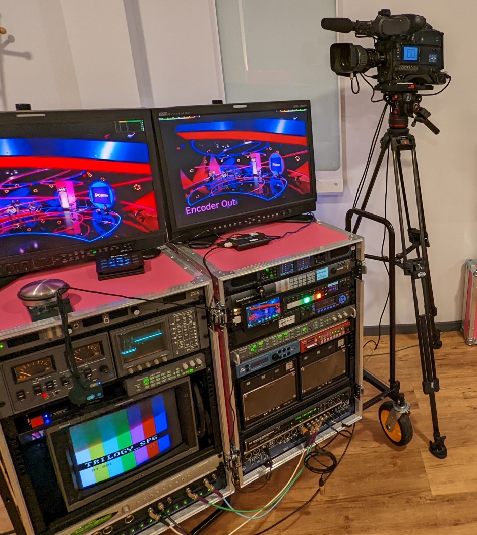 2 portable 19 inch equipment racks full of broadcast demonstration hardware with 2 broadcast SDI monitors on top and an ENG camera on a tripod beside them.