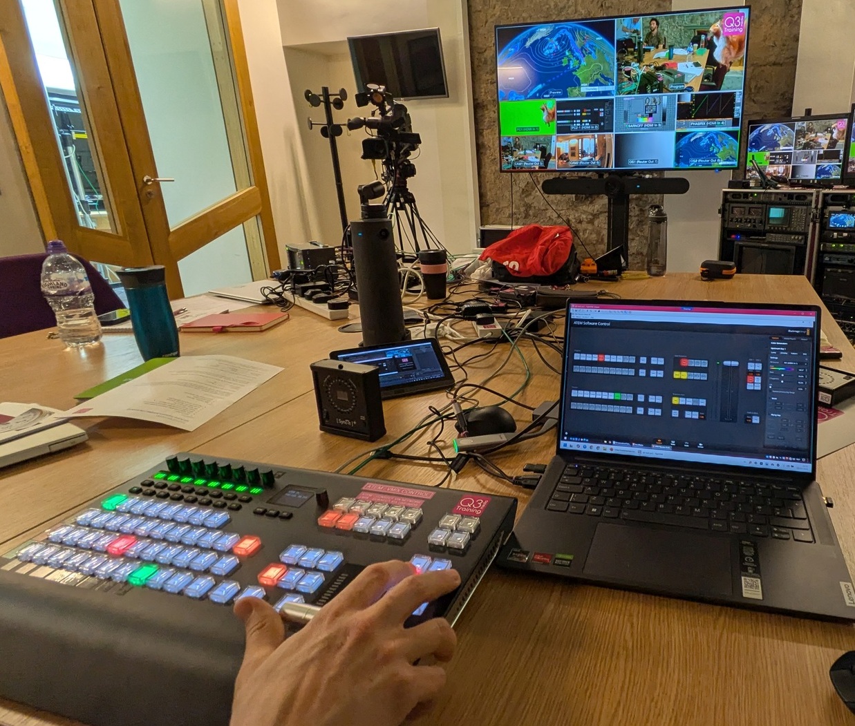 Practical Kit - Vision Mixer 2 View across table where a vision mixer control panl and control software on a laptop can be seen with a large multiviewer display in the background.